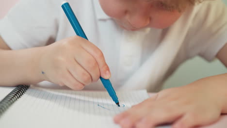 little boy draws on notebook page with blue marker at table