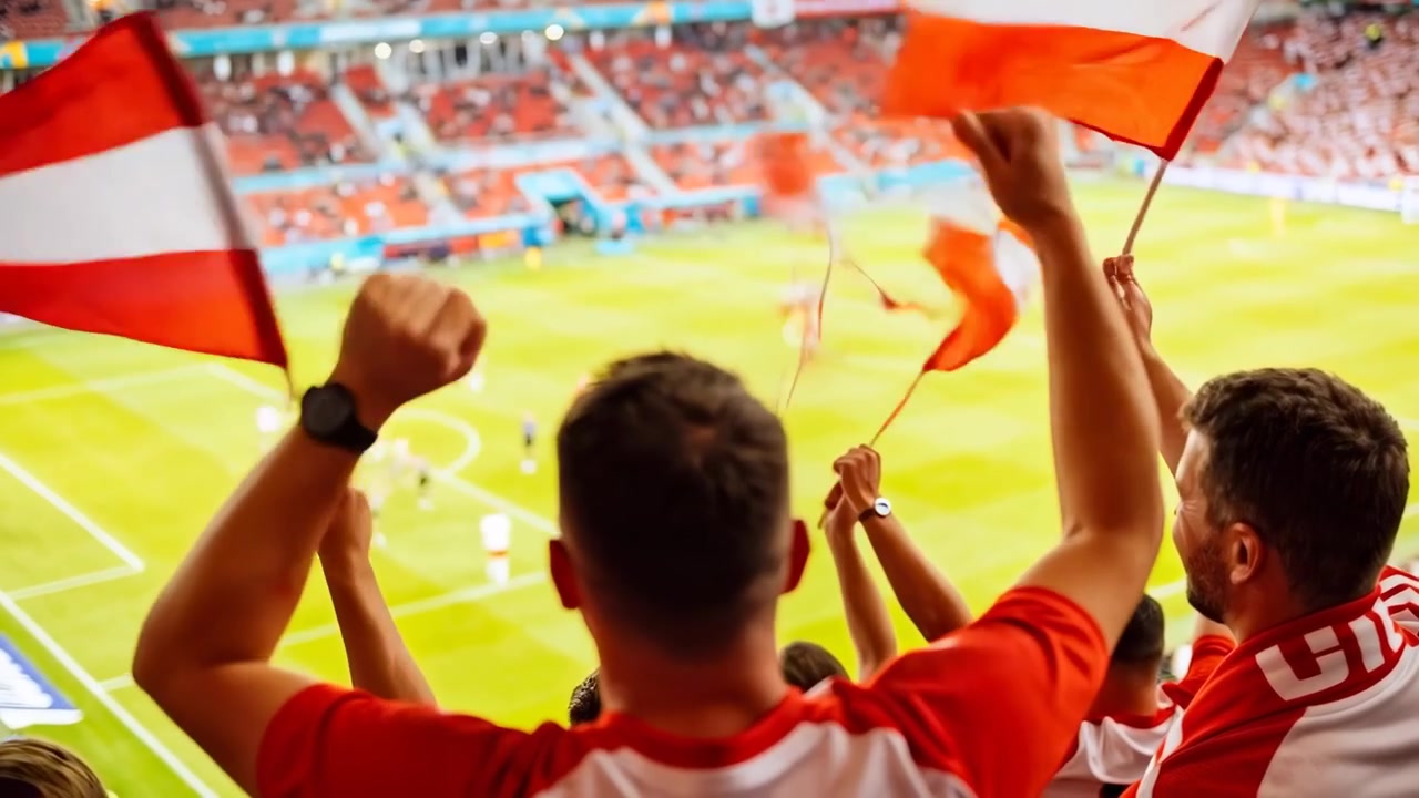Reagindo ao jogo em andamento no estádio, torcedor de camisa vermelha no centro levantando os braços e agitando a bandeira, torcendo.