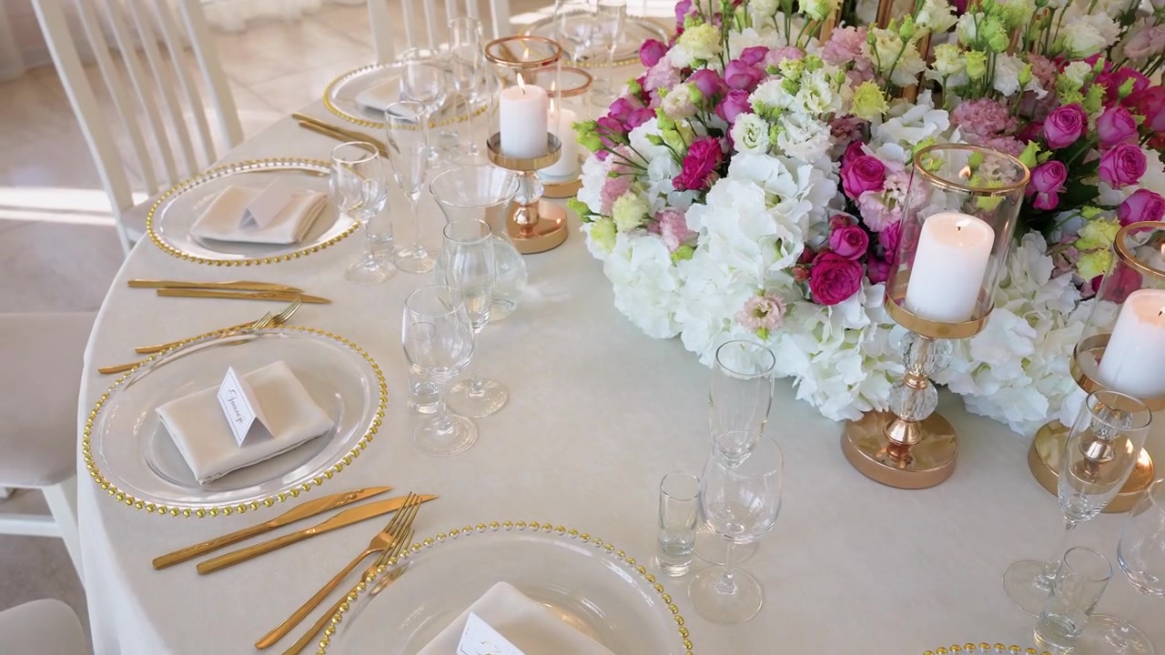 Luxurious wedding reception table setting featuring a lavish floral centerpiece, glowing candles, gold cutlery, and glassware, all bathed in soft natural daylight. No people present.