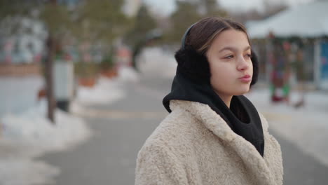 young lady in warm beige winter coat and black hoodie turns around with light smile on snowy street, soft blurred background with decorated trees, snow-covered ground, and cozy winter atmosphere