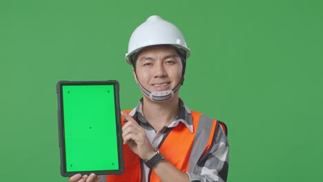 Close Up Of Asian Male Engineer With Safety Helmet Smiling And Showing Green Screen Tablet To The Camera While Standing In The Green Screen Background Studio