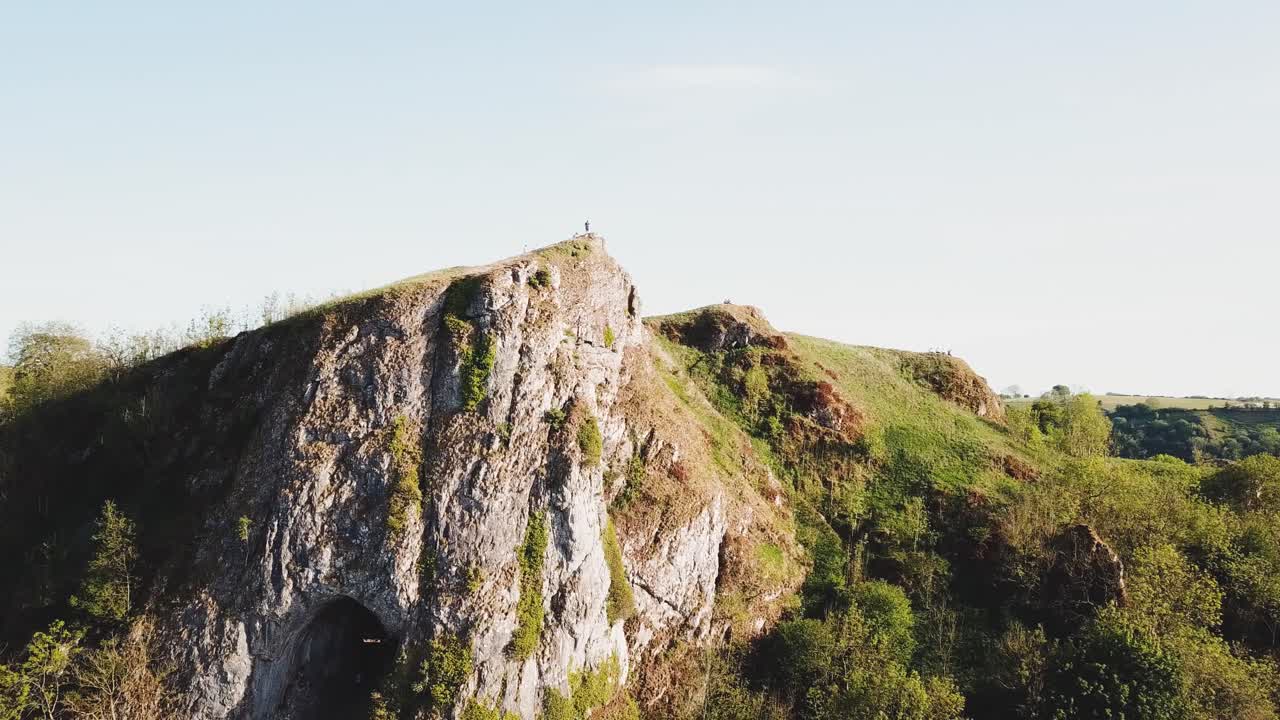 Clockwise aerial drone shot featuring Thor's Cave, Ashbourne, Peak District, England. Large rock formations, green hills and clear blue skies.