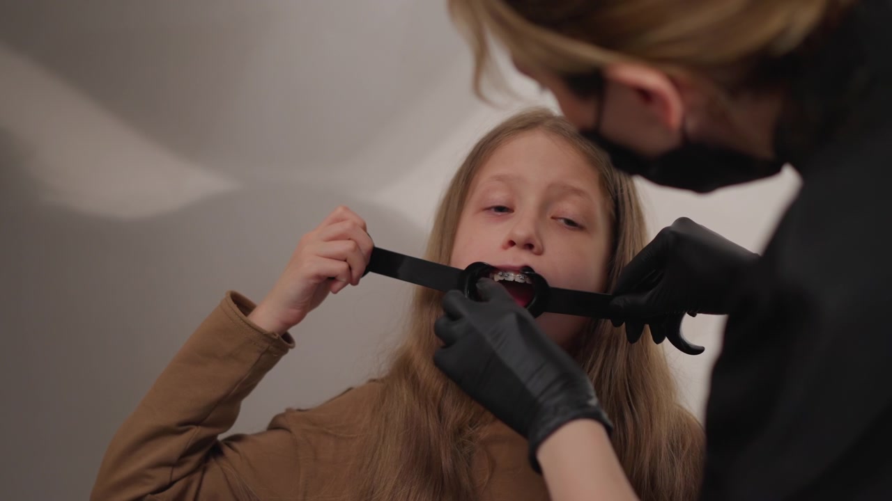 Primo piano di una giovane ragazza che tiene la bocca aperta mentre l'ortodontista, con guanti neri, esamina e regola l'apparecchio dentale usando attrezzature specializzate. La scena presenta un'illuminazione soffusa con messa a fuoco