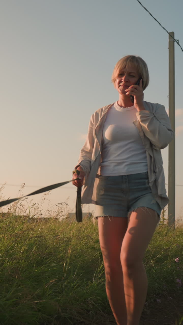 Dog owner in jean shorts on phone call walking along grassy field at dawn, holding leash while dog pulls forward energetically, electric pole and distant building visible in background
