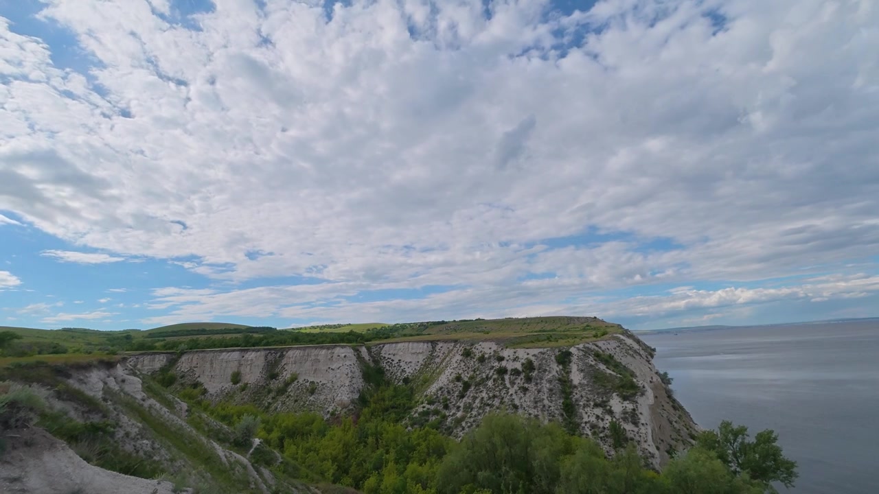 Et time-lapse viser hvide kridtklinter dækket af grønne træer med udsigt over en bred flod under en dynamisk blå himmel med hurtigtbevægende hvide skyer.