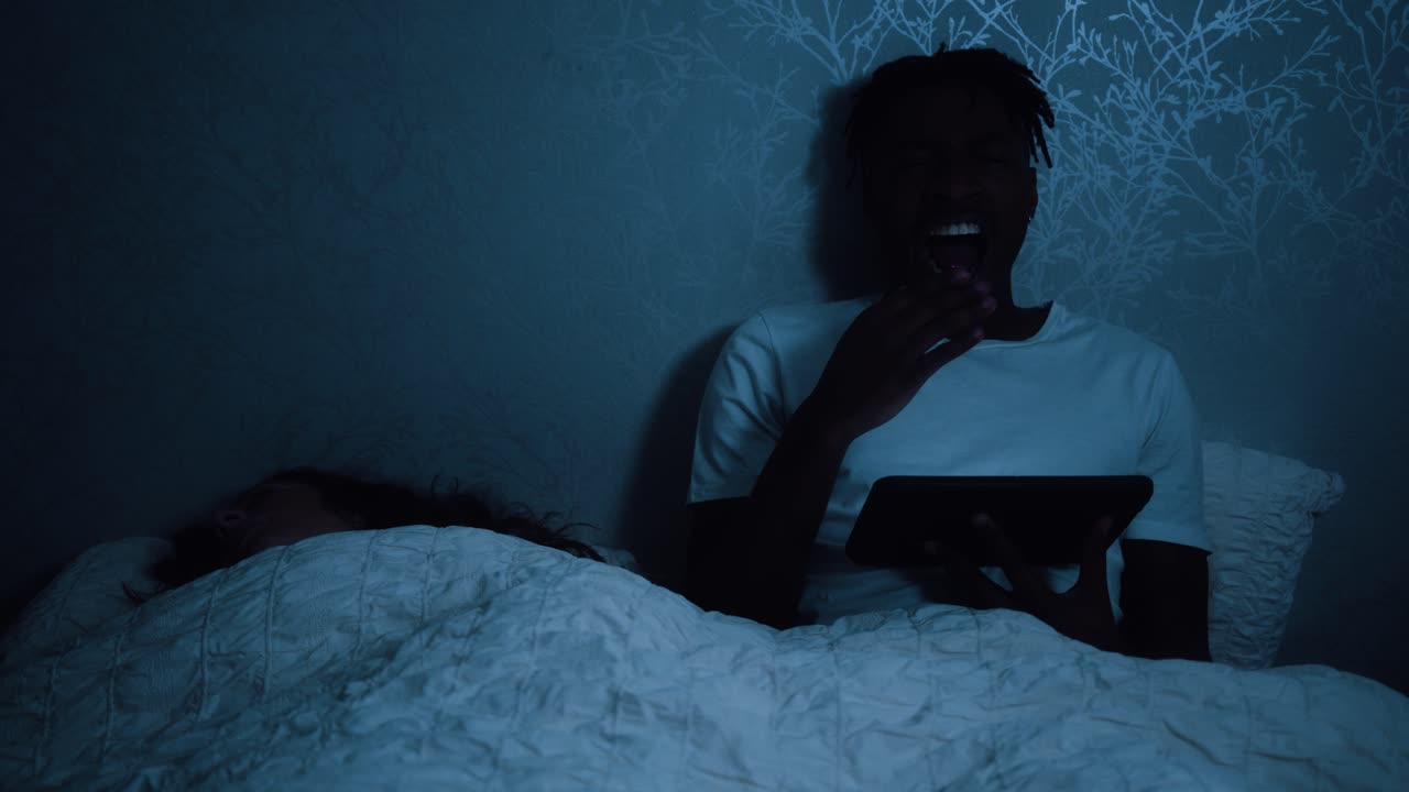 Man yawns whilst sat in bed at night watching a tablet device
