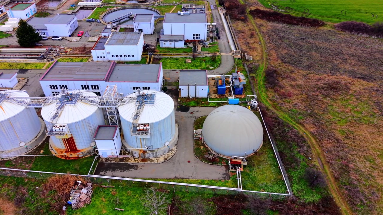 Elevated aerial view of wastewater treatment tanks, Bulgaria. Cylindrical digester towers, dome gasholder and white facility buildings with green surroundings.