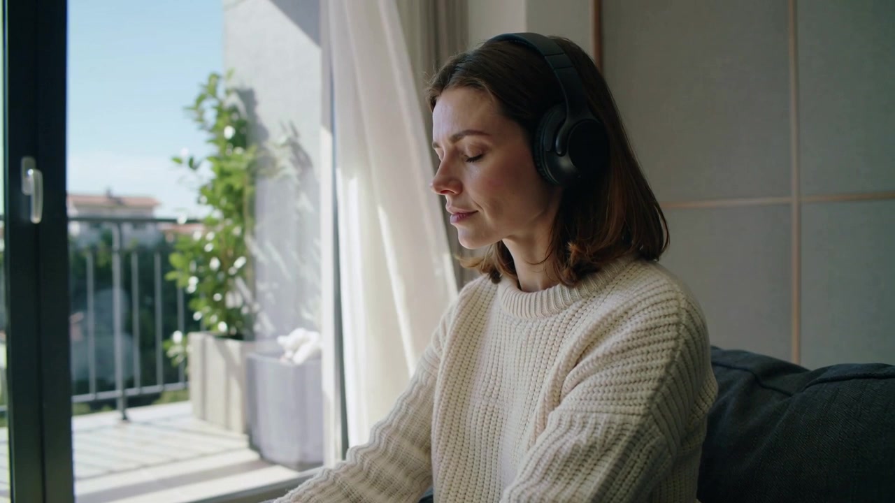 Con gli occhi chiusi, una donna con le cuffie medita per rilassarsi sul divano vicino al balcone, indossando un maglione color crema.