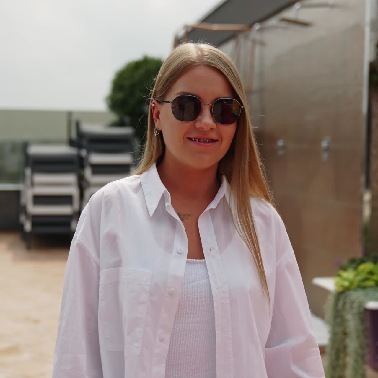 Beautiful long-haired young lady wearing white swimsuit and shirt walks slowly outdoors. Blonde woman wearing sunglasses smiles to the camera