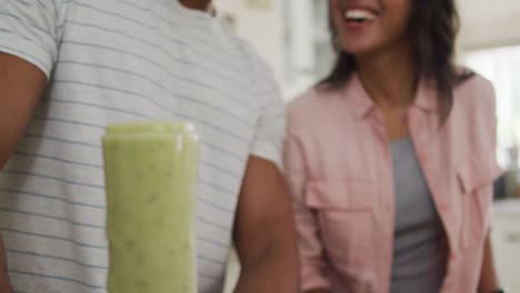 close up of happy biracial couple in kitchen preparing smoothie, using blender