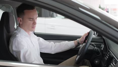 A Young Buyer in a White Shirt is Sitting in the Interior of a New Car. A young buyer in a white shirt is sitting in the interior of a new car. A man of European appearance inspects the car from the inside