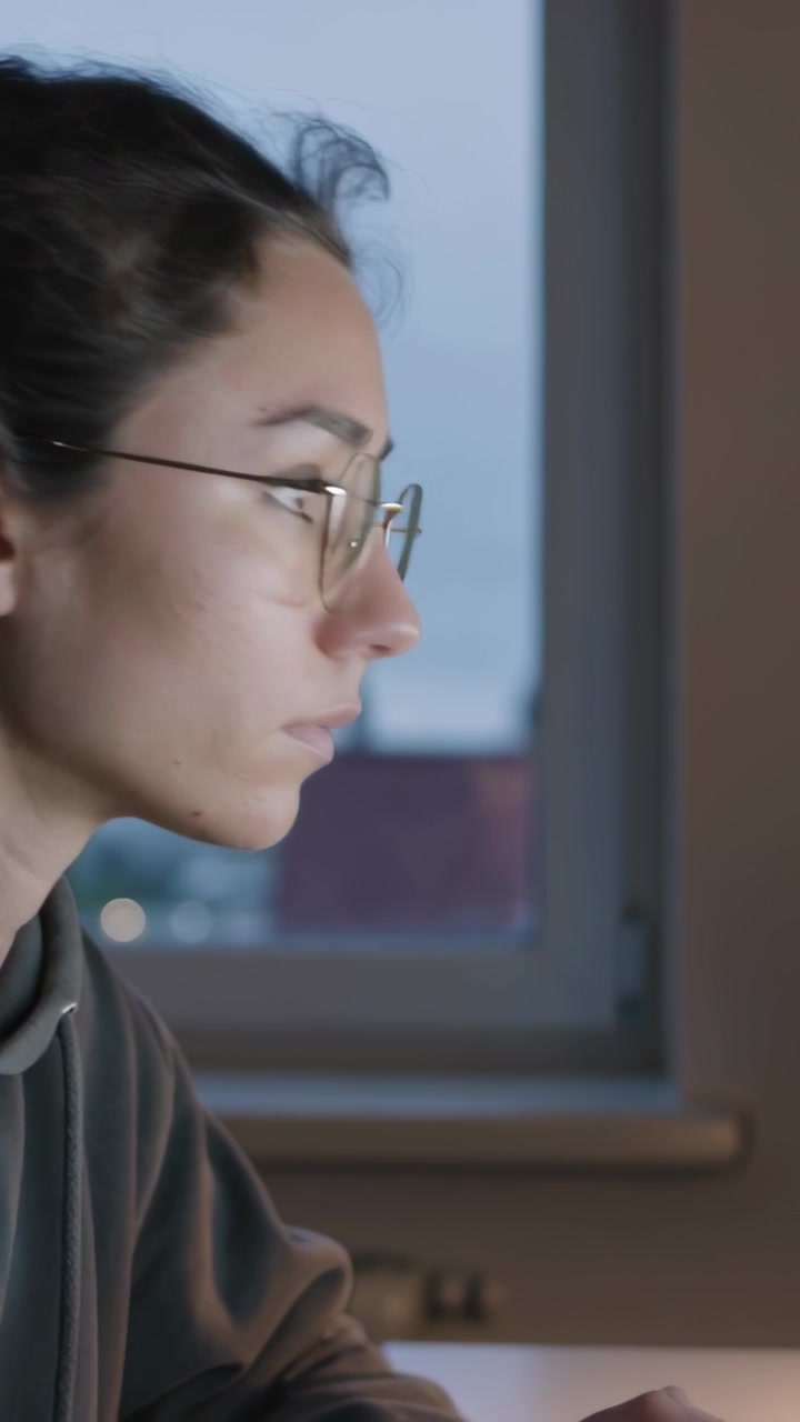 Video vertical: Reaccionando a la pantalla, mujer inclinada cambiando la mirada en un escritorio junto a la ventana, con gafas y sudadera con capucha.