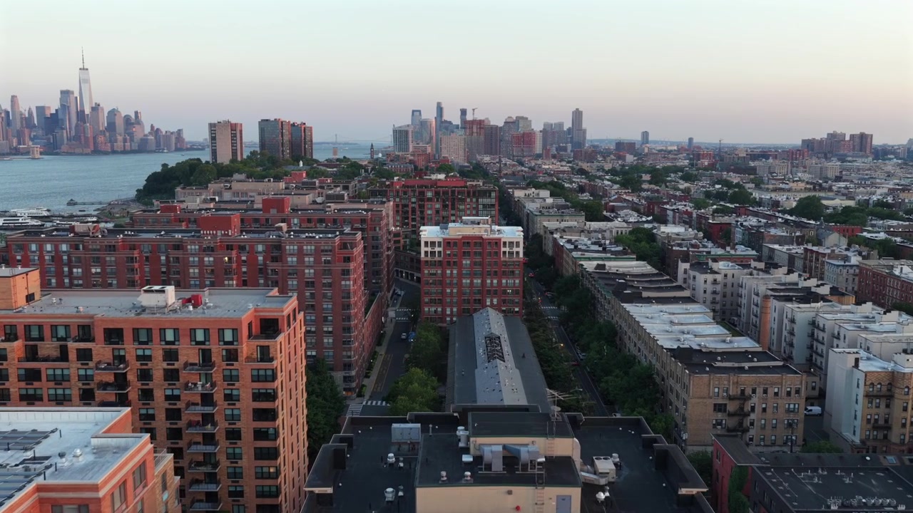 Røde murstenslejlighedsbygninger fylder et tæt bebygget havnefrontkvarter i Hoboken, New Jersey. One World Trade Center rejser sig på den anden side af den rolige Hudson River under en diset gylden solnedgang. Droneoverflyvning.