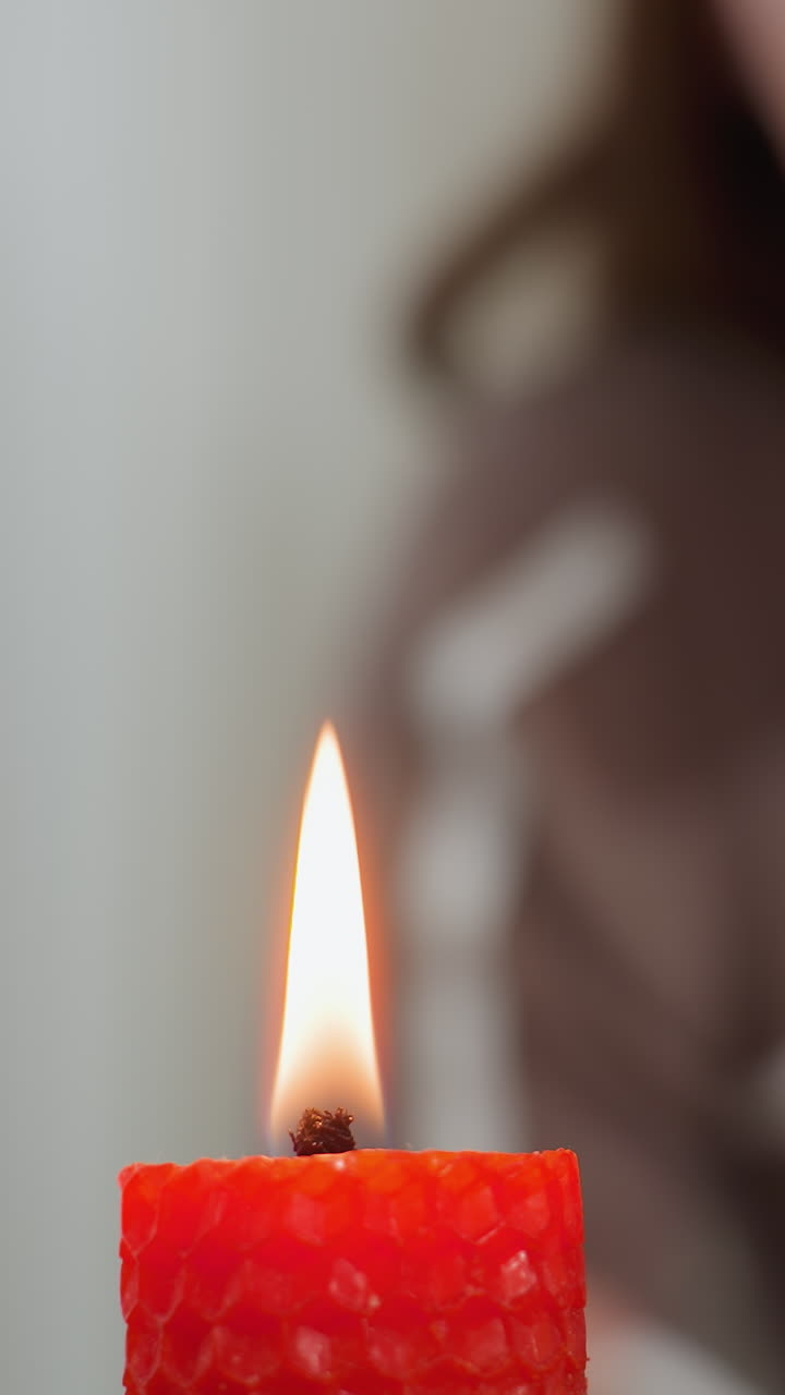 Primo piano di una fiamma di candela rossa mentre qualcuno sullo sfondo si prepara a spegnerla; la messa a fuoco morbida sulla persona aggiunge profondità, mentre il bagliore caldo della fiamma contrasta con l'ambiente circostante sfocato