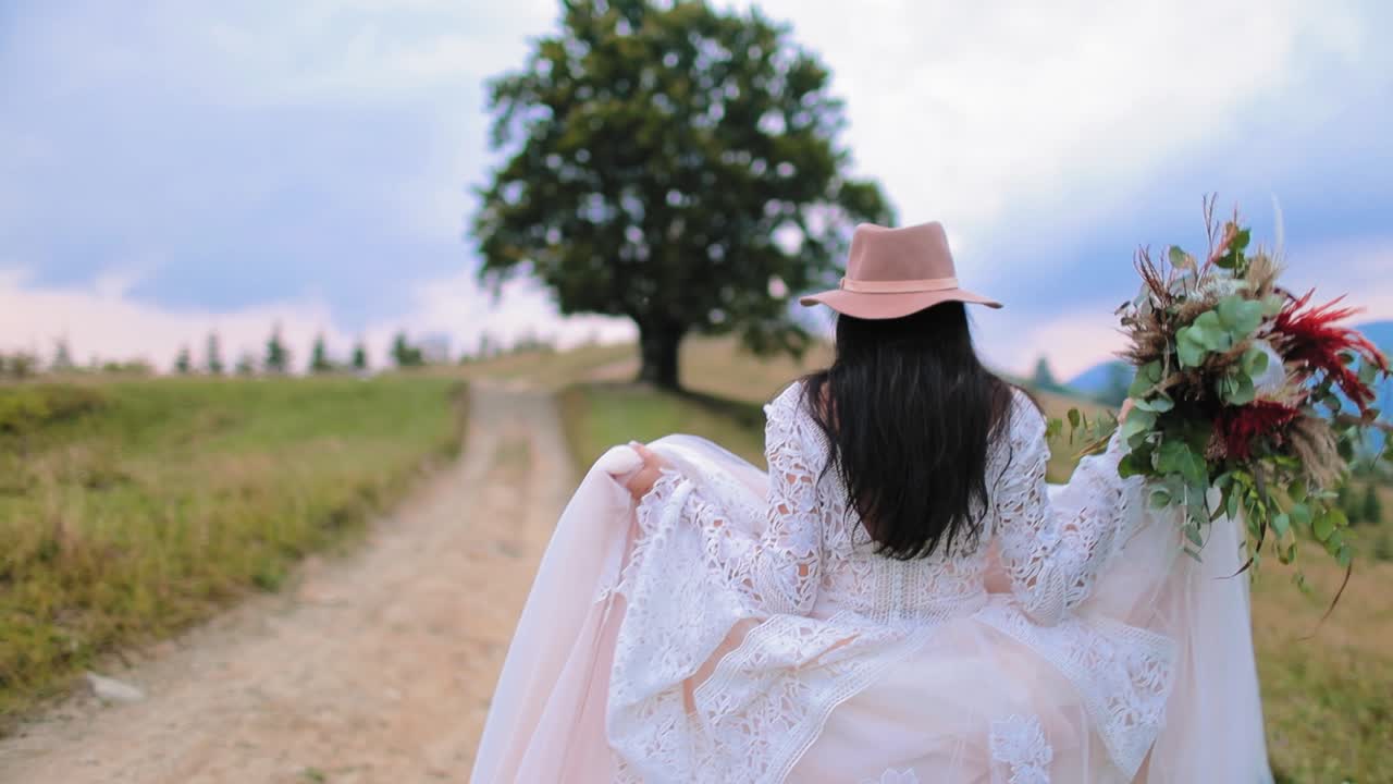 Bride walking in nature. Backside view of attractive girl in luxury wedding dress and pink hat in the mountains landscape. Slow motion.