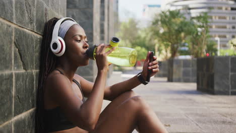 african american woman exercising outdoors drinking water and using smartphone in the city