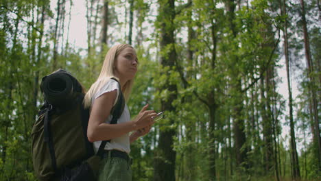 a young woman with a mobile phone walks through the forest traveling with a backpack in slow motion. traveler in shorts in the woods looking for gps satellites