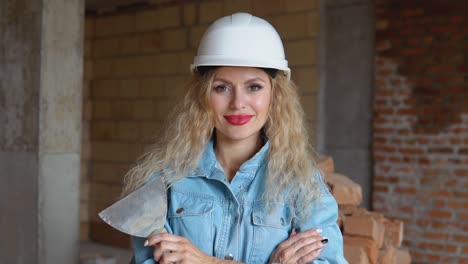 a female builder in a helmet and denim work uniform stands on a construction site. young woman bricklayer with makeup and manicure holding a spatula
