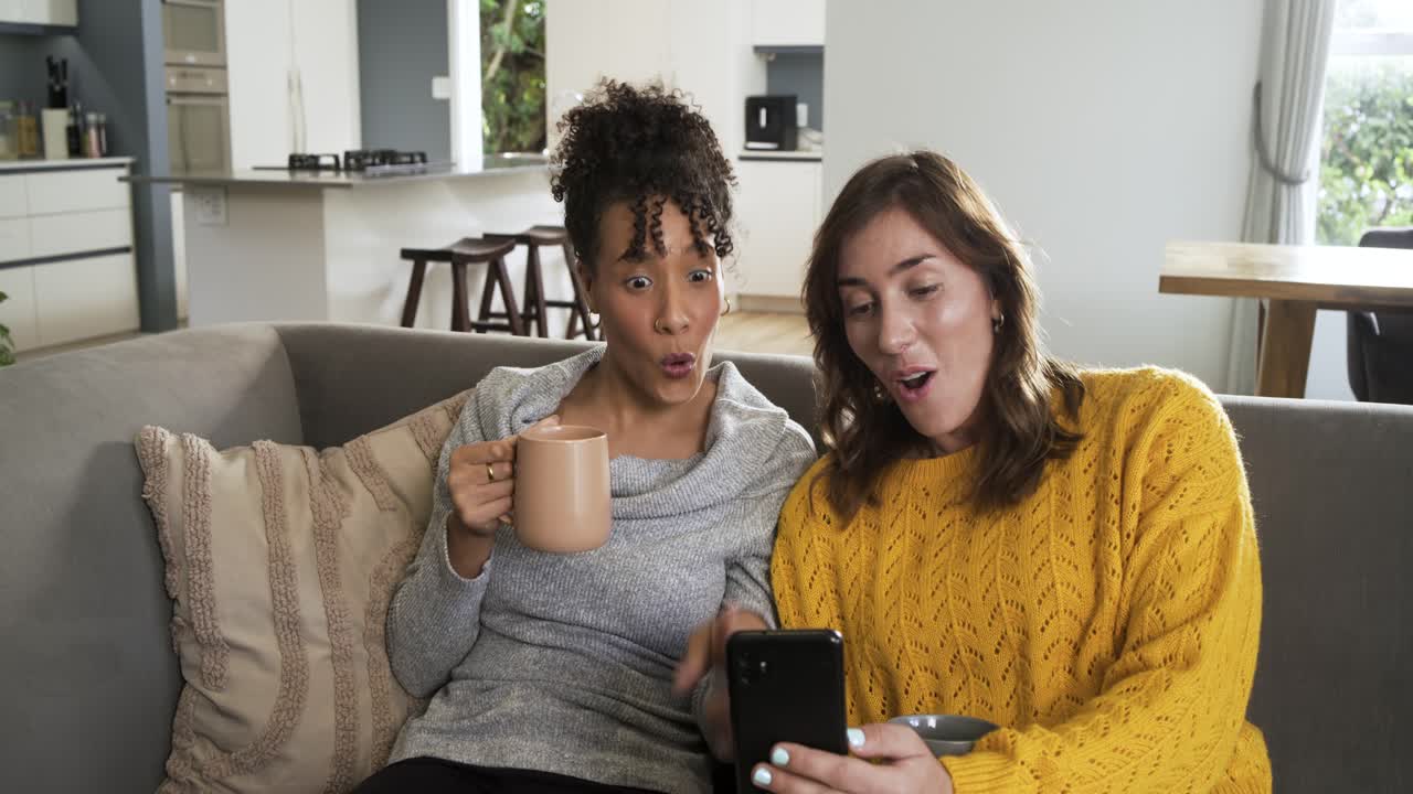 Amigos de meia-idade no sofá, com canecas, notam mensagem surpreendente no smartphone e compartilham risadas.