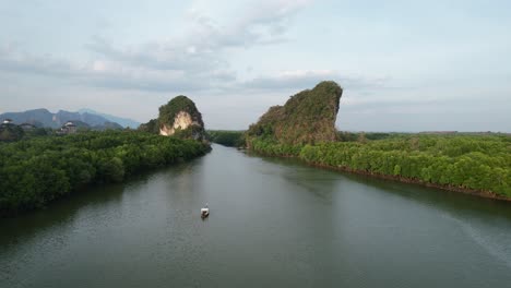 drone aéreo volando hacia atrás revelando un bote de cola larga tailandés en un río entre dos grandes montañas de piedra caliza verde y bosque de manglares en la ciudad de krabi tailandia al atardecer