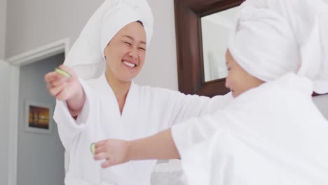 video of happy asian mother and daughter in robes doing cucumber mask and having fun