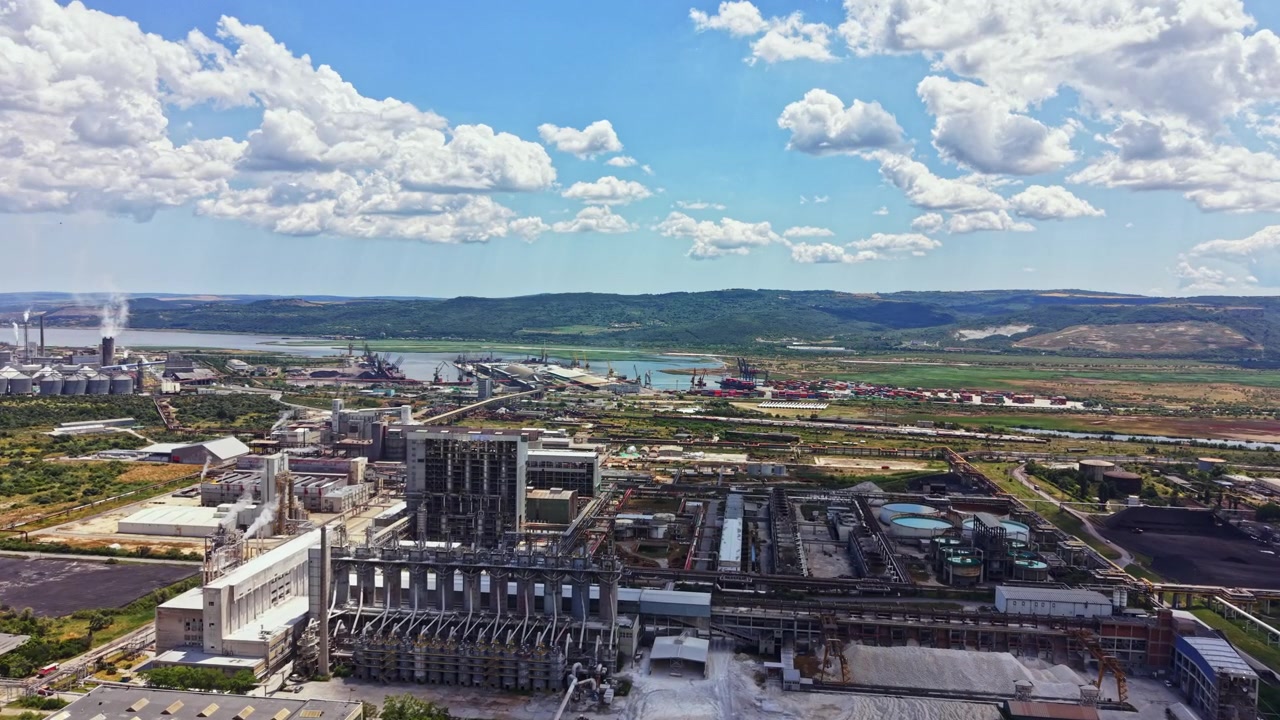 Vista di un'area industriale vicino a un fiume con fabbriche e macchinari sotto un cielo nuvoloso in una giornata di sole.