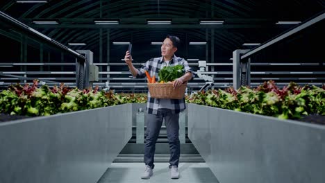 cuerpo lleno de granjero masculino asiático con canasta de verduras teniendo una llamada de videoconferencia con alguien a través de teléfono inteligente en la granja inteligente