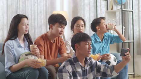 asian teenagers cheering, eating and watching football game on smart phone at home