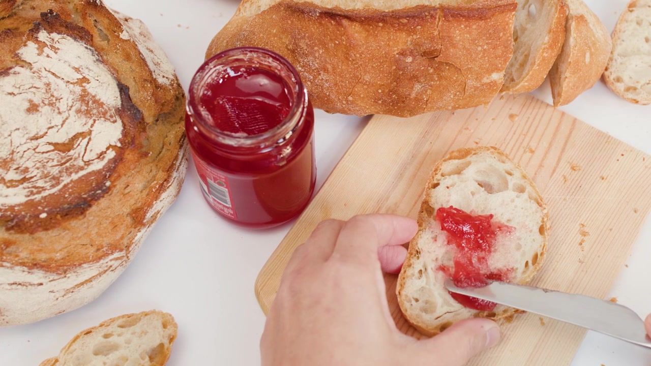Overhead view of hands spreading red fruit jam on crusty bread using a metal knife.