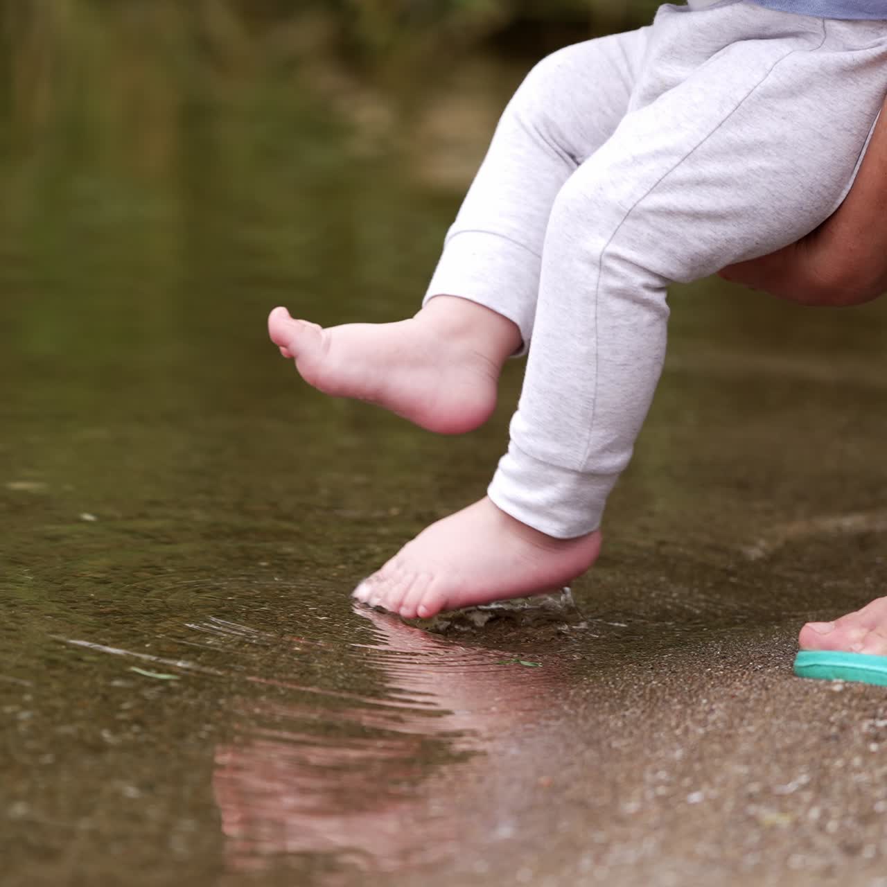 Tiny baby feet trying water in the pond. Mom's hands holding a baby close to the river and kid deeps them into water