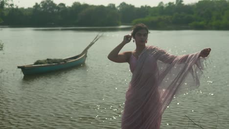 woman in pink saree by the river, adjusting her attire, with a traditional boat in the background, daylight