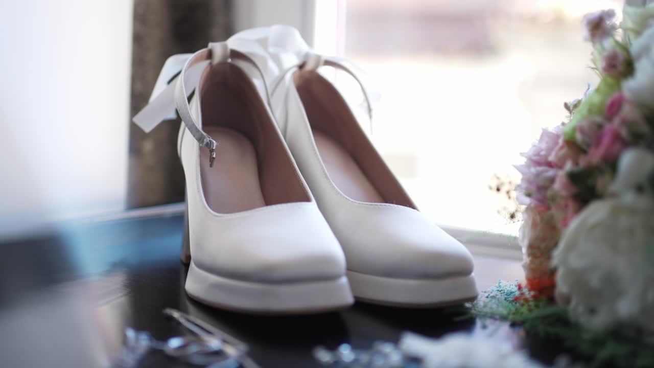 White wedding shoes and a bridal bouquet rest near a window in soft natural light