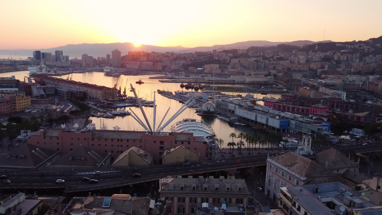 Drone flies backward and slightly upward over Genoa historic port at sunset, showing traffic, city, and mountains in the background