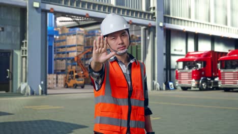 asian male engineer with safety helmet disapproving with no hand sign while standing , outside of logistics distributions warehouse