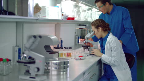 Mujer-Científica-Mirando-Microscopio.-Asistente-De-Laboratorio-Lleva-Libreta-A-Mujer-Científica