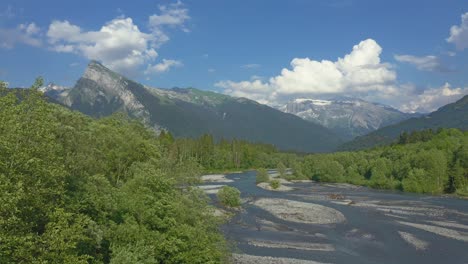 Aerial-view-above-the-Giffre-River-in-the-French-Alps,-flying-above-the-beautiful-vibrant-green-spring-trees-with-a-beautiful-mountain-view-of-the-Criou-in-the-background