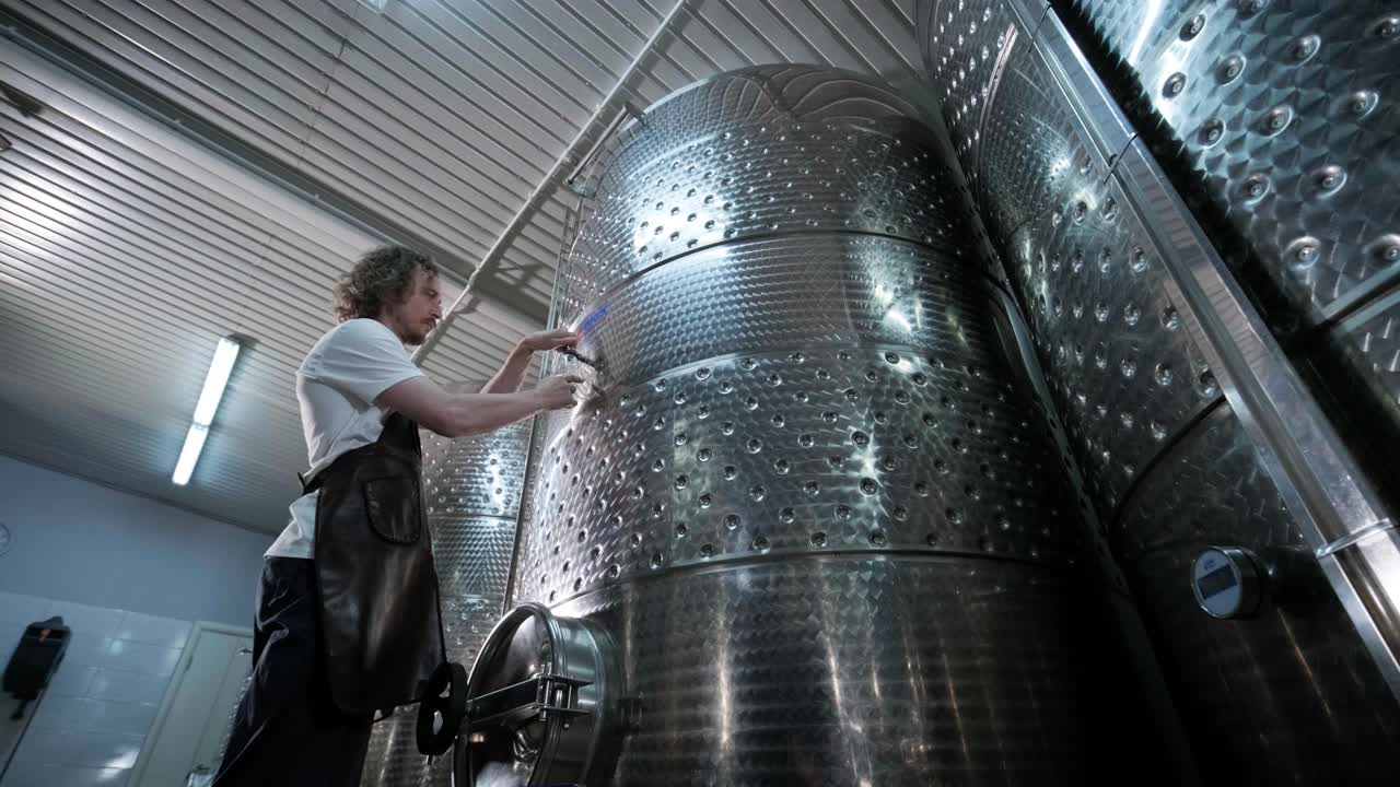 un agricultor o viticultor feliz está caminando en medio de tanques de vino y comprobando un grado de fermentación de la producción de vino de alta calidad en una instalación moderna de elaboración de vino o bodega.