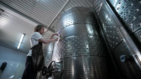 a happy successful farmer or winemaker is walking in the middle of wine tanks and checking a fermentation grade of high quality wine production in a modern winemaking facility or winery.