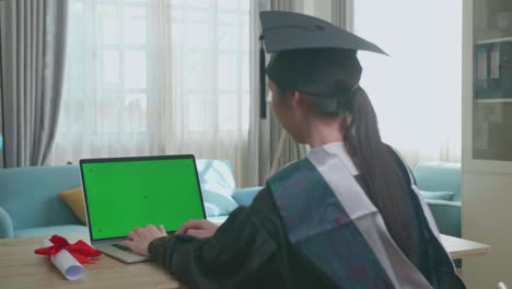 asian woman wearing a graduation gown and cap use laptop computer with green mock-up screen in living room