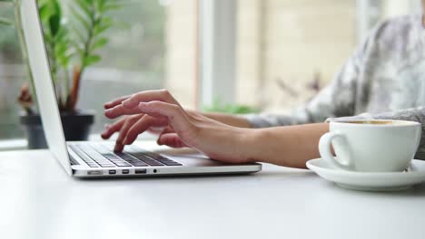 Vista-De-Cerca-De-Las-Manos-De-Una-Mujer-Trabajando-En-Una-Computadora-Portátil.-La-Mujer-Está-Sentada-Cerca-De-La-Ventana-En-La-Cafetería-Y-Escribiendo.-Taza-Blanca-Con