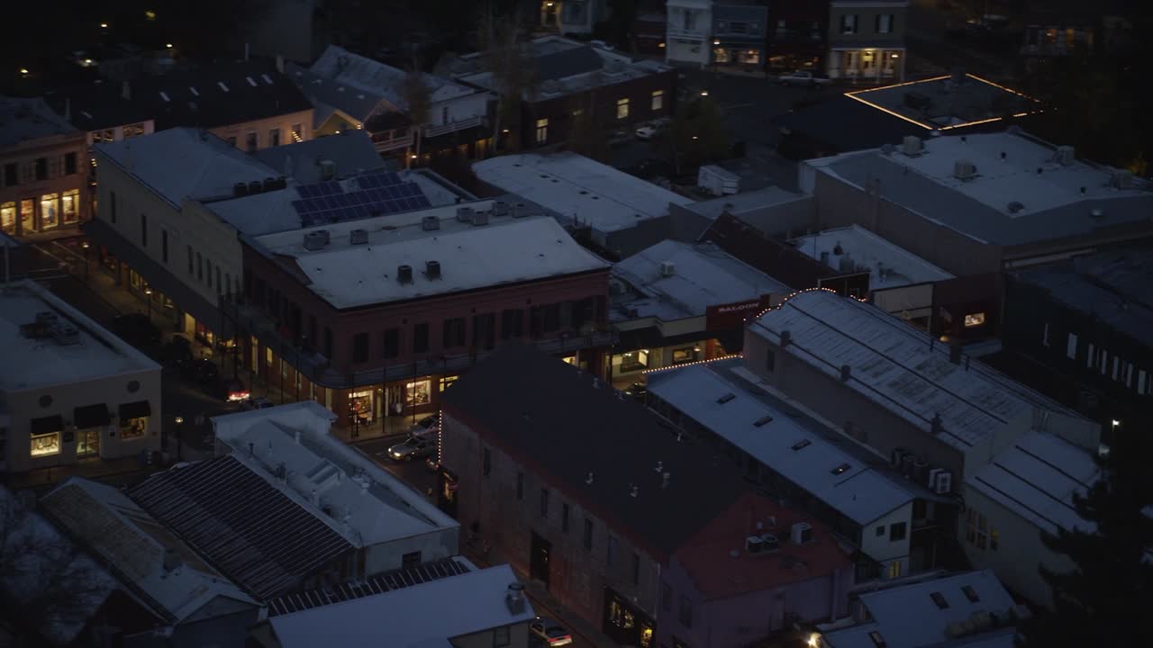 Footage of a small urban area at dusk, illuminated by streetlights and glowing interiors. The aerial view showcases diverse architectural styles with flat and gabled rooftops, some equipped