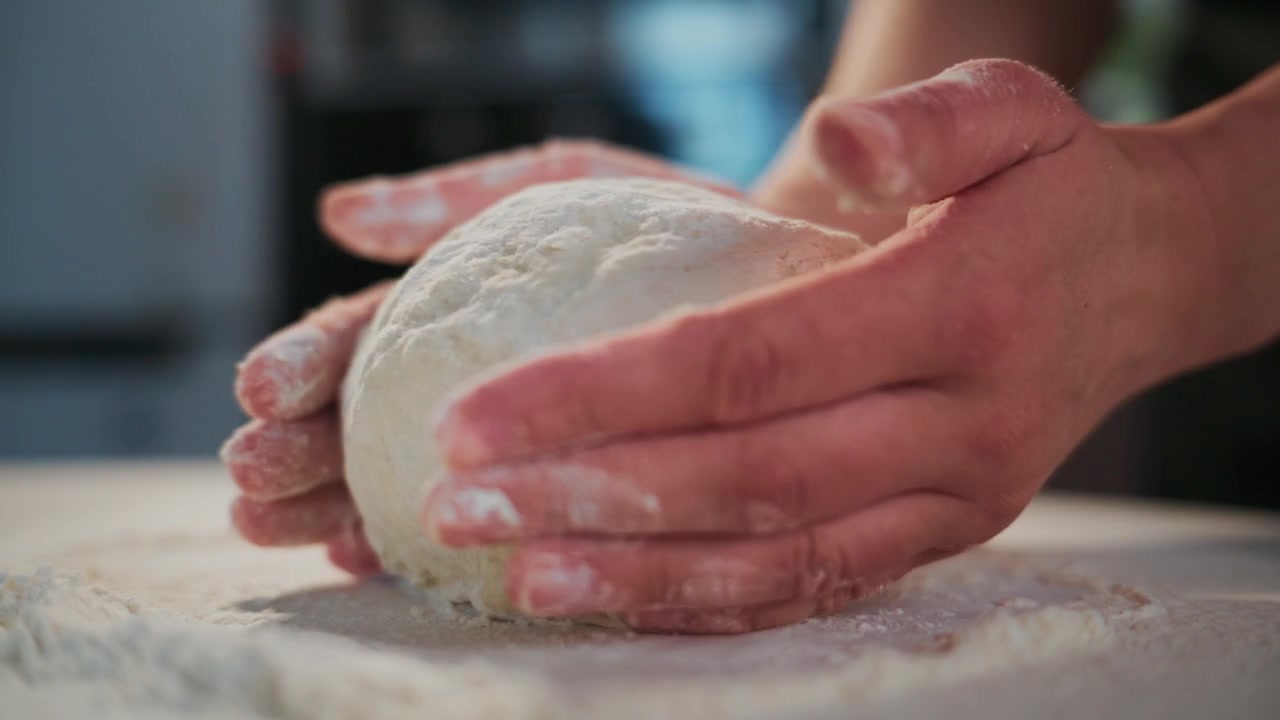 Le mani impastano un impasto morbido su una superficie infarinata. Focus sull'azione tattile e sulla consistenza. La scena mostra una preparazione del cibo calma e metodica. Ideale per ricette, tutorial di cottura o contenuti culinari