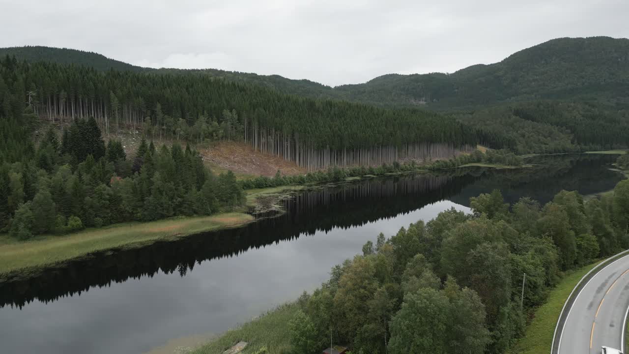 Flying over road towards the extremely calm and still lake showing reflections of the green lush trees forest in Norway with