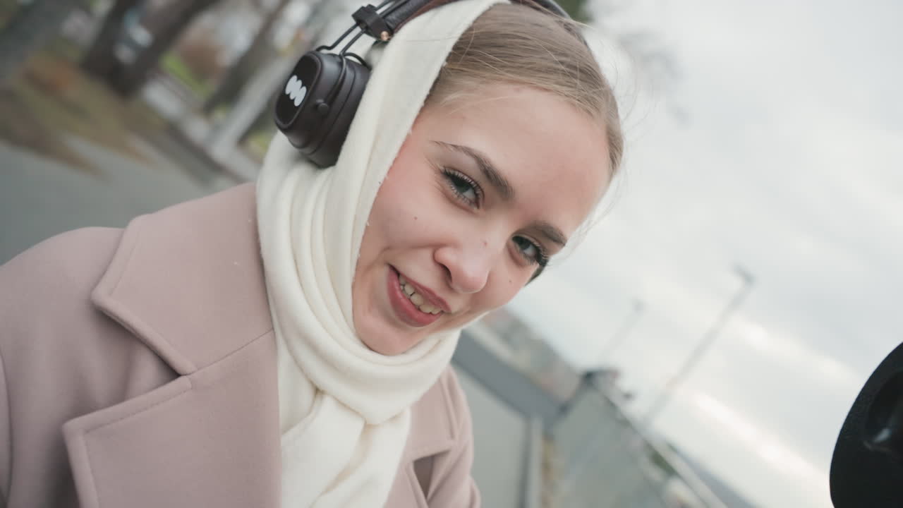 Donna in cuffie al belvedere cittadino osserva attraverso un telescopio pubblico e fa il gesto delle corna, sorride con sciarpa e cappotto beige, sfondo di lungomare, energia da vlog spontaneo, atmosfera urbana invernale.