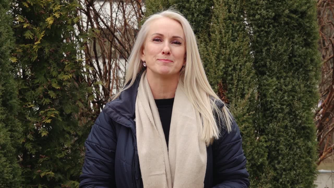 A woman with long blonde hair smiles and speaks while standing outdoors in front of evergreen trees, dressed warmly in a navy coat and beige scarf.