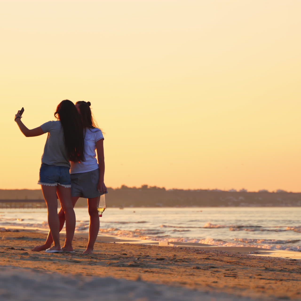 Young women posing to camera on the phone at the seaside at sunset. Two girls taking selfie in mobile phone in the evening sea.