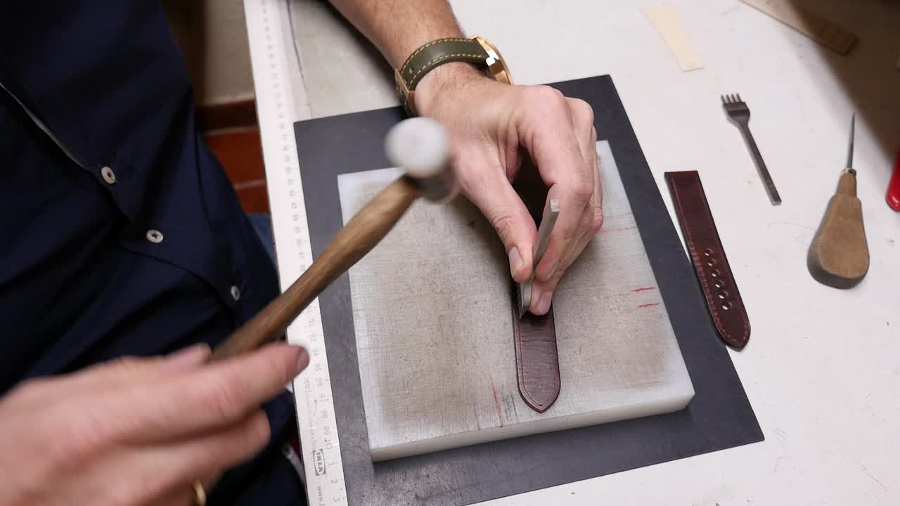 The hands of an artisan hammering and shaping the leather of a strap on a wooden workbench. The manufacturing process of a traditional, handmade leather accessory