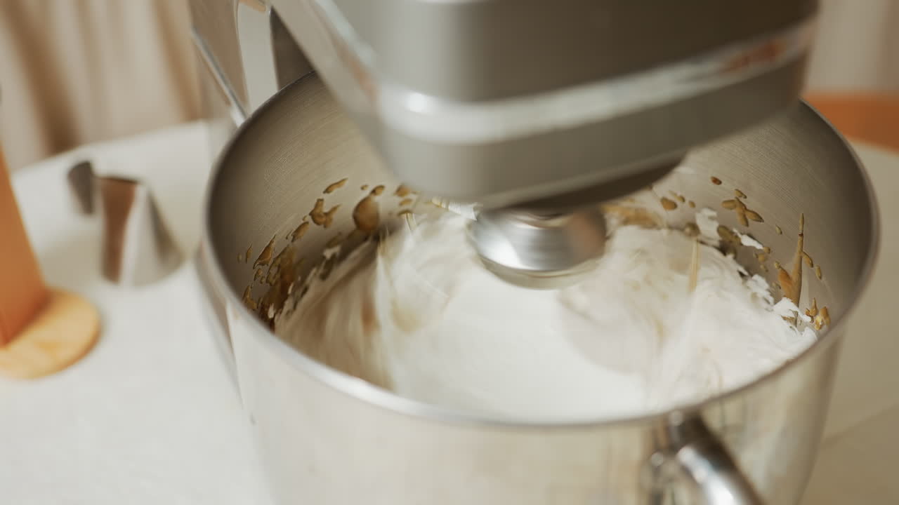 Close up of electric stand mixer blending fluffy white batter with visible brown splashes around stainless steel bowl, surrounded by baking tools on kitchen table during baking preparation process