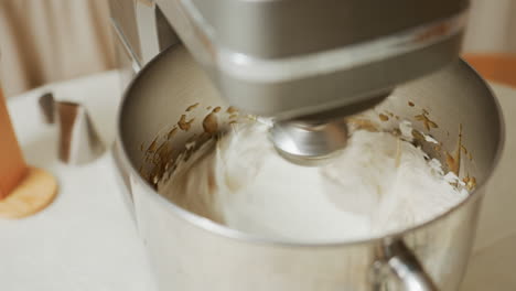 close up of electric stand mixer blending fluffy white batter with visible brown splashes around stainless steel bowl, surrounded by baking tools on kitchen table during baking preparation process
