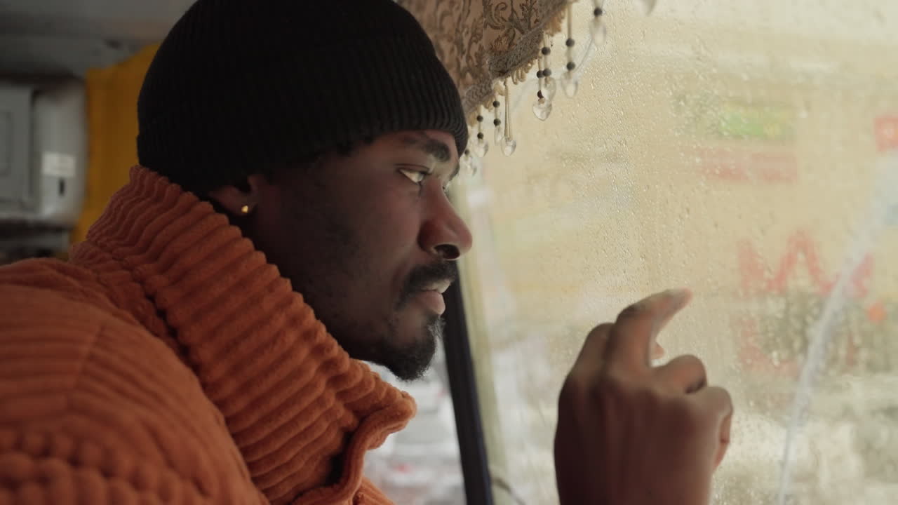 Black man taxi driver looking out through rainspeckled window, knit beanie and ribbed orange sweater, raindrops on glass, urban street blurred outside, contemplative gaze, hand tapping glass, interior